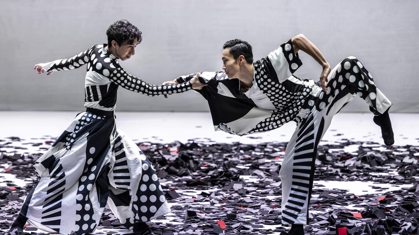Två dansare, båda iförda lösa kläder tryckta med geometriska mönster, dansar på en scen beströdd med små svarta rutor.