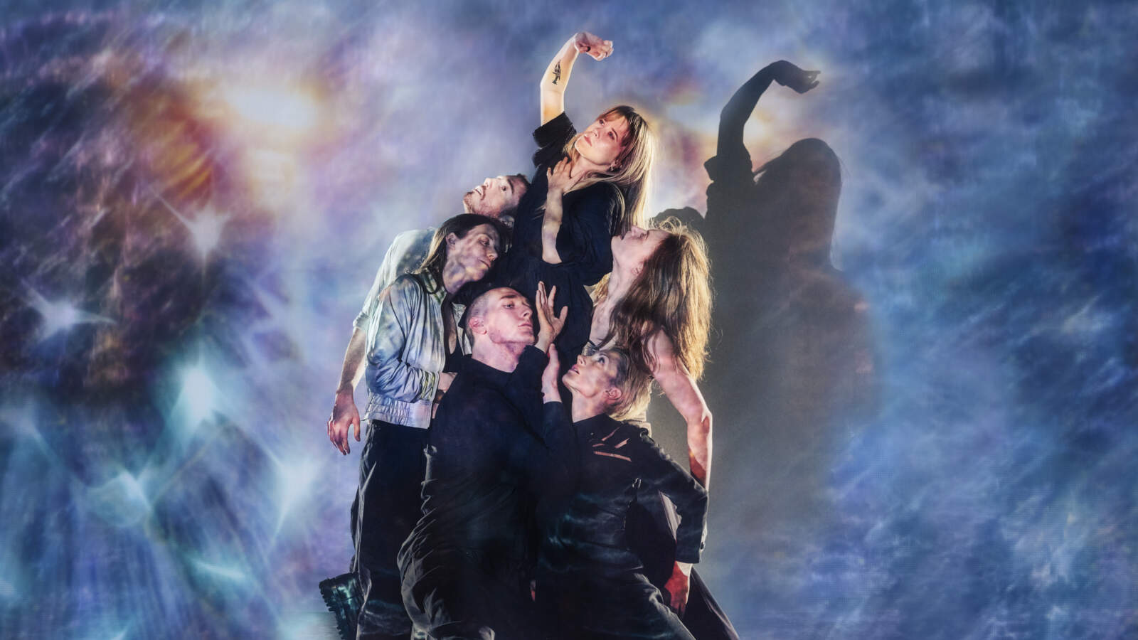 Sex dansare står tätt runt Anna som höjs över dem med en arm sträckt uppåt. Ljus projiceras över gruppen och fonden bakom.