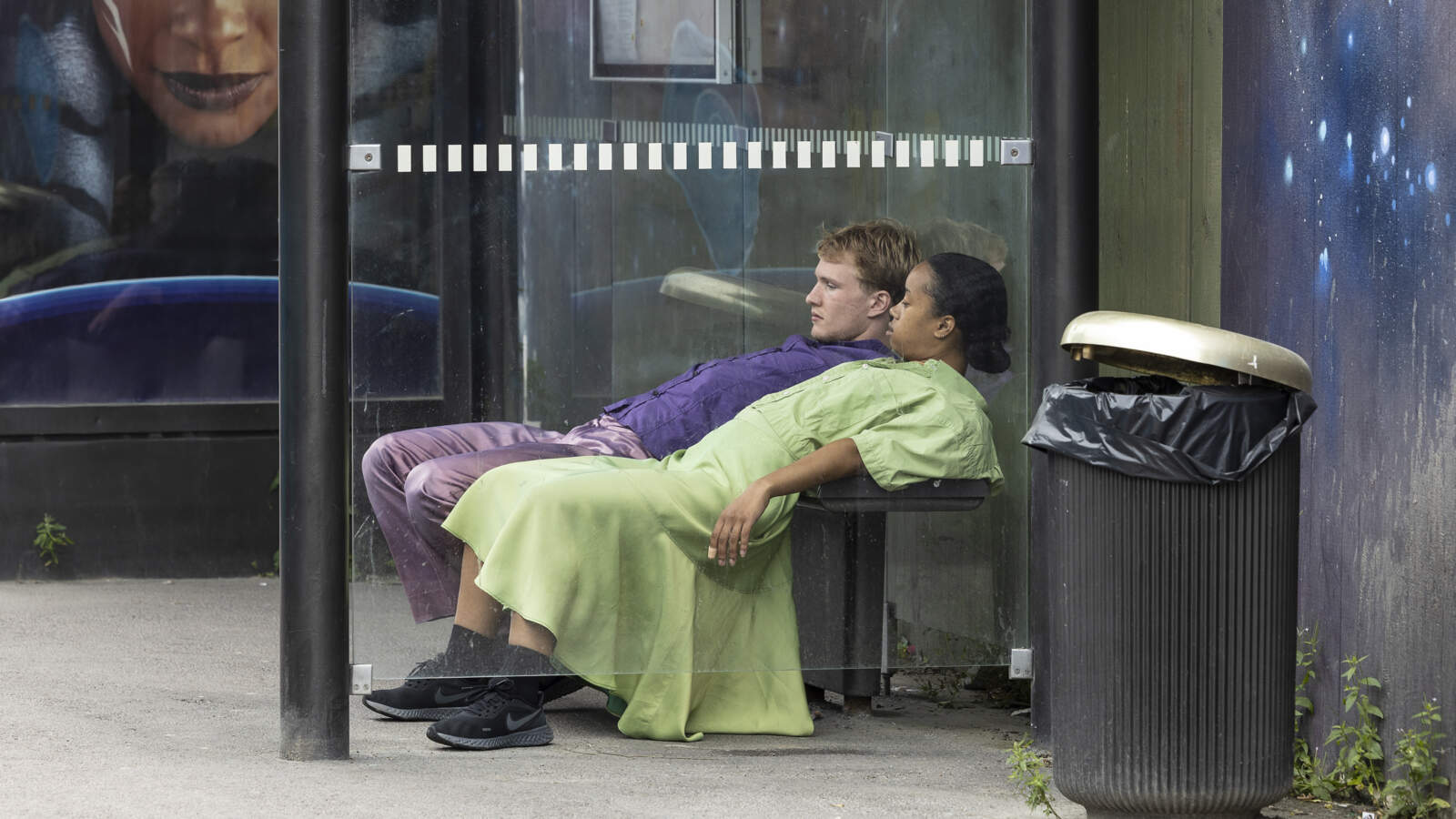Två dansare vilar bara överkroppen mot en bänk i ett busskur, benen böjda framför dem och höfterna upplyfta.