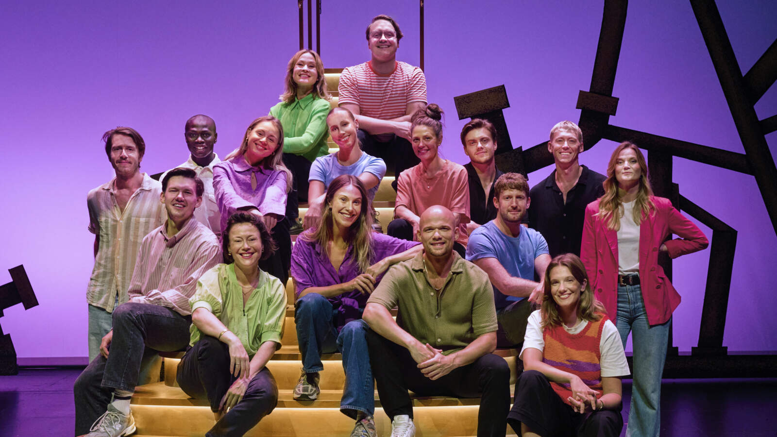 A group of musical artists sit on a golden staircase on a stage in front of a purple backdrop. They are smiling.