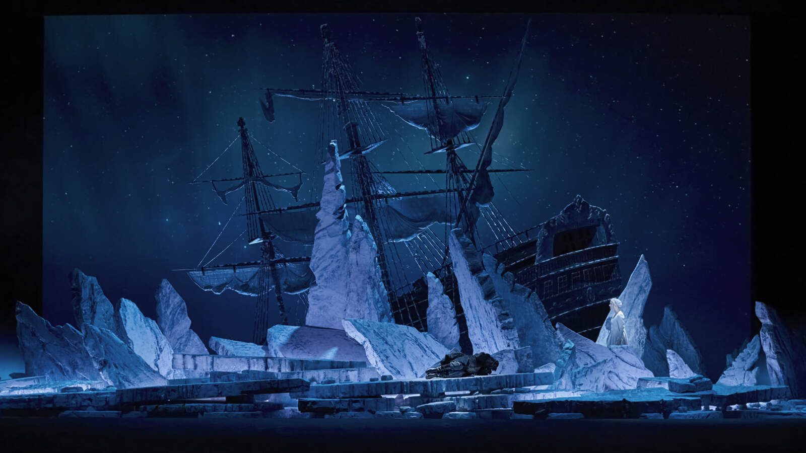 Ett stort segelfartyg står fastbunden bland isflak som pekar i olika riktningar. Någon ligger på isen i fosterställning.