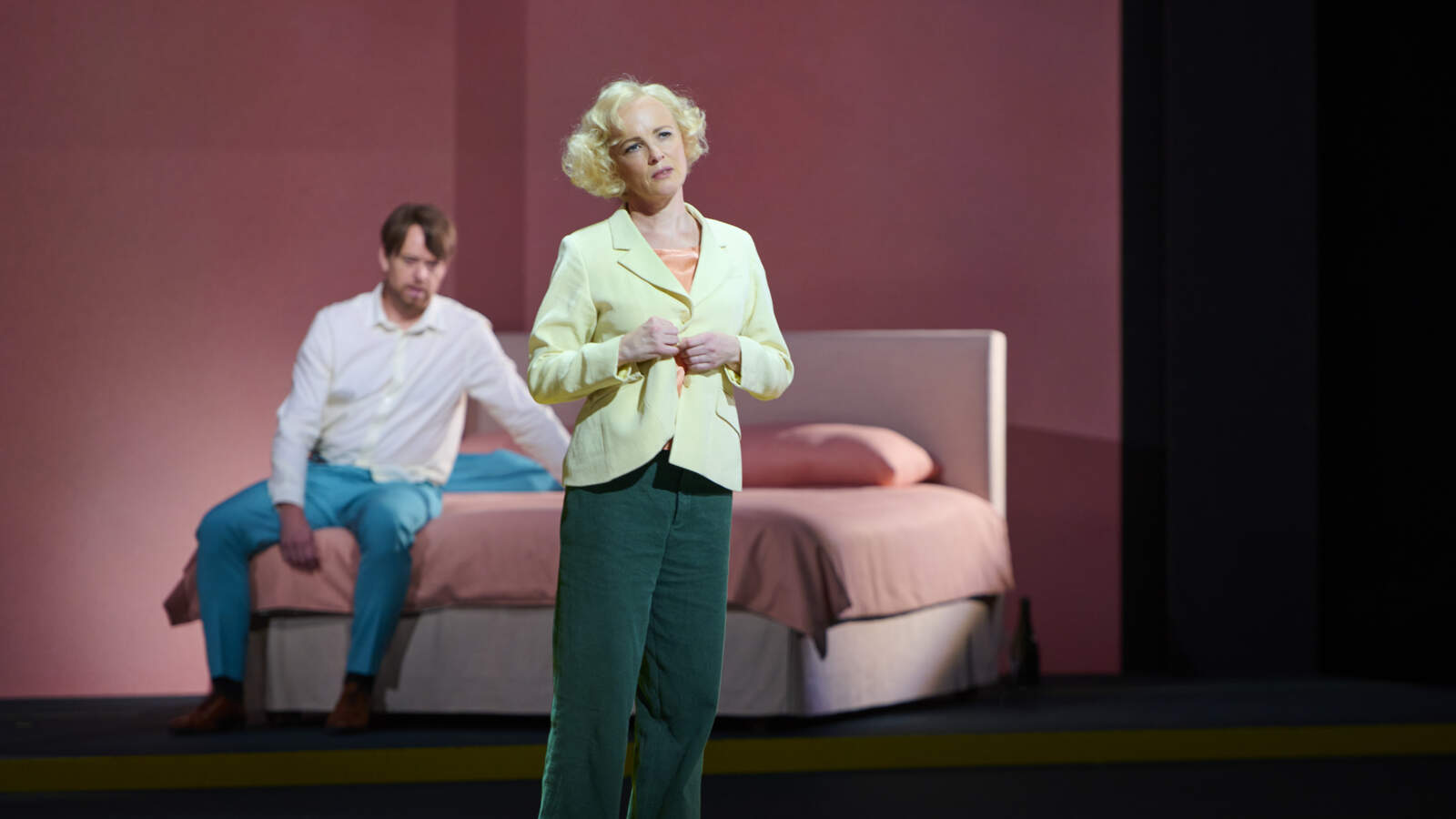 Nelly står i ett rum med rosa väggar och knäpper upp sin jacka. Francis sitter bakom henne på en säng.