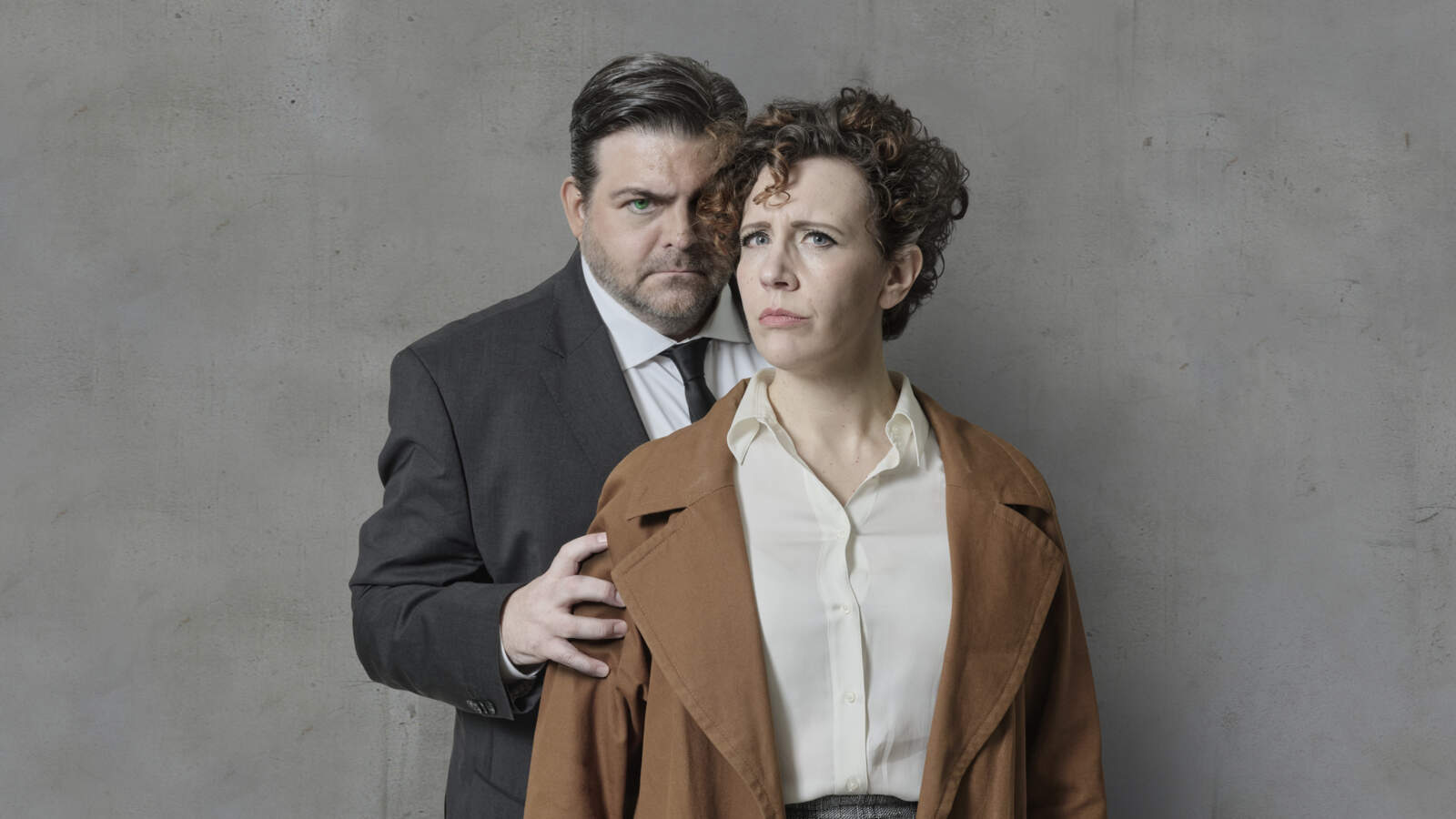 En ilsken man och en orolig kvinna står framför en betongvägg. Mannen håller kvinnans axel. Båda tittar mot betraktaren.
