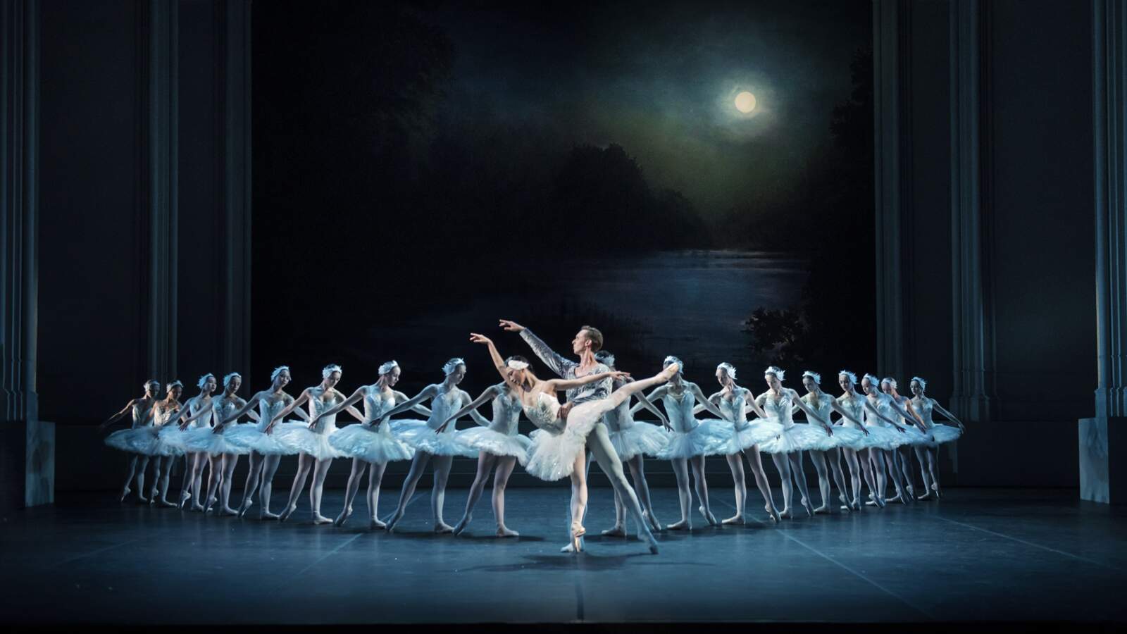 Siegfried och Odette dansar på en mörk scen, framför en rad vitklädda ballerinor. Bakom dem lyser en fullmåne.