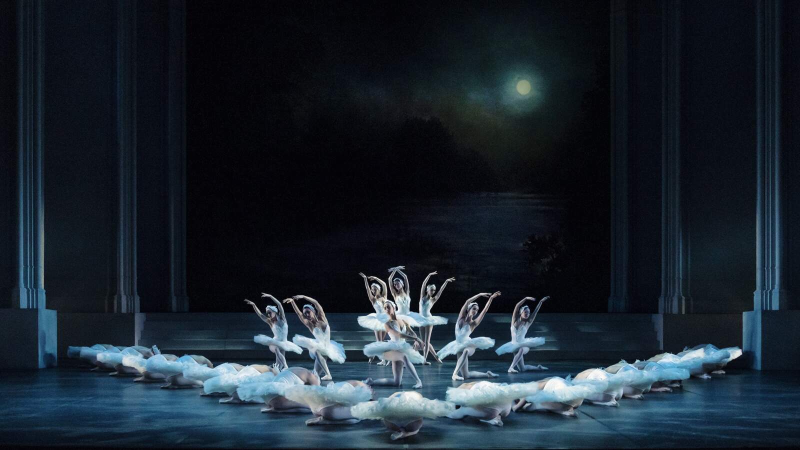 En grupp vitklädda ballerinor dansar synkroniserat på en mörk scen som svanar på en sjö. En fullmåne lyser bakom dem.