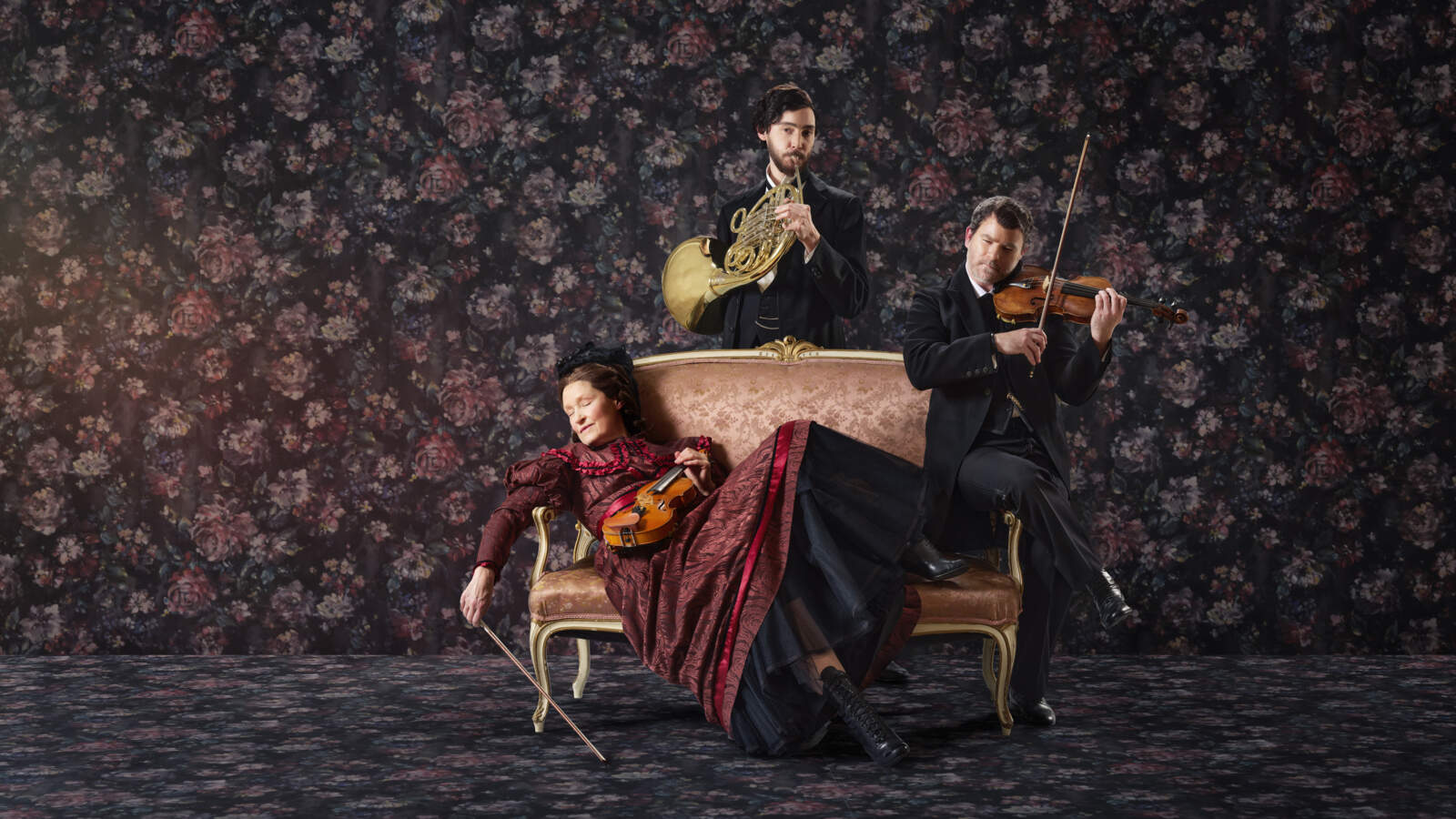 En violinist ligger med slutna ögon på en schäslong i ett tapetserat rum, medan en annan violinist samt hornist spelar.