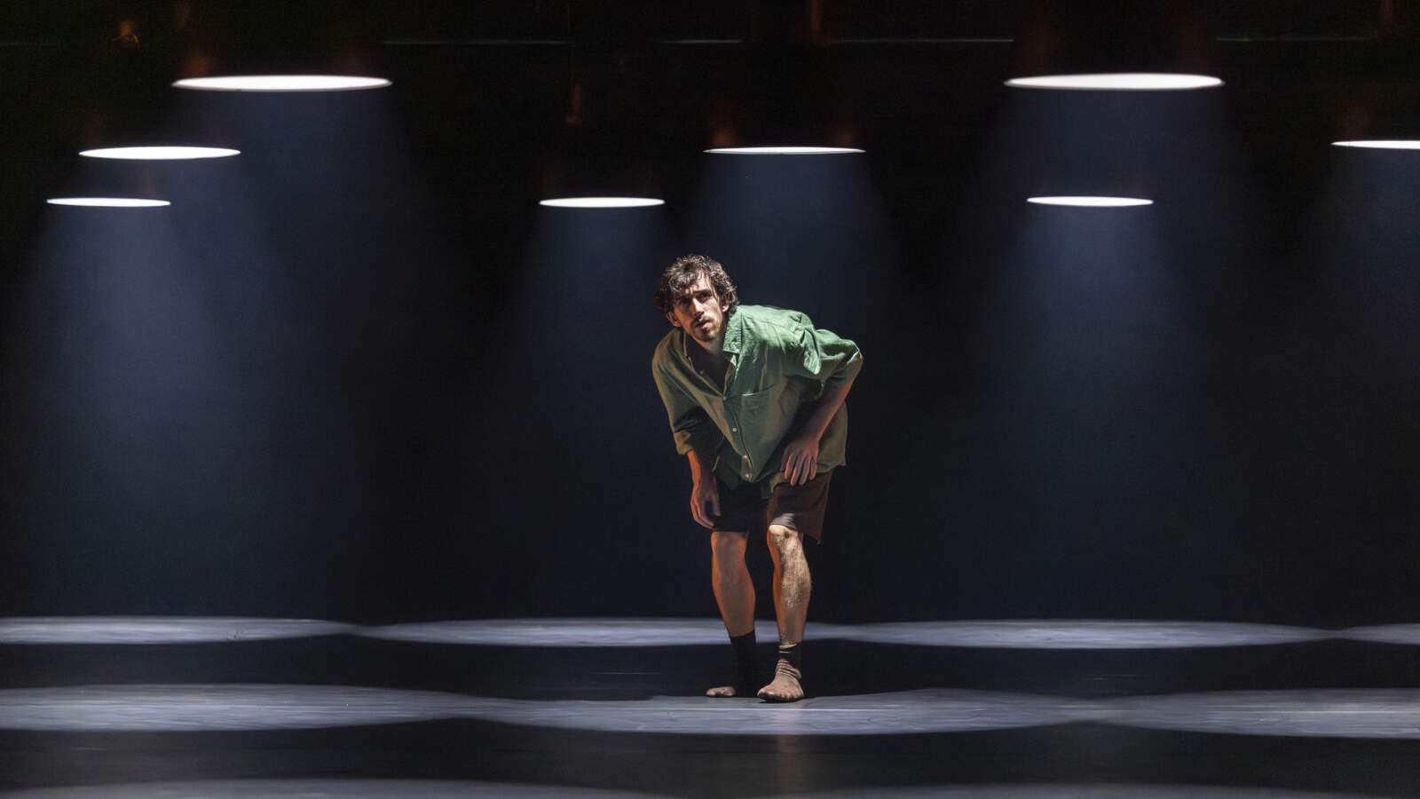En dansare lutar sig framåt och tittar upp. Han står under en ljusrigg med spotlights som hänger lågt ovanför honom.