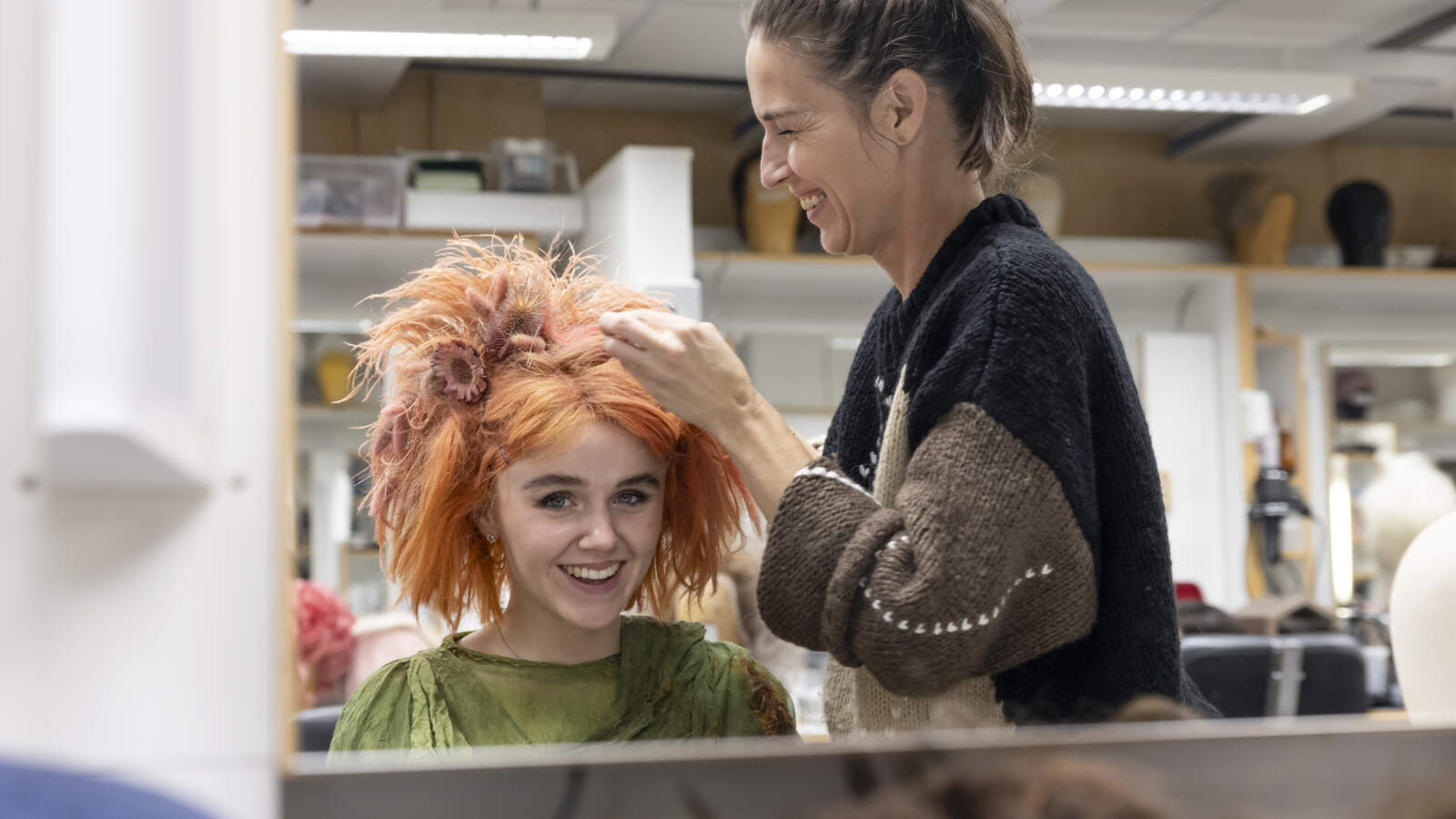 En perukmakare tittar på den röda peruken som bärs av en skådespelare som sitter i en stol. Båda ler.