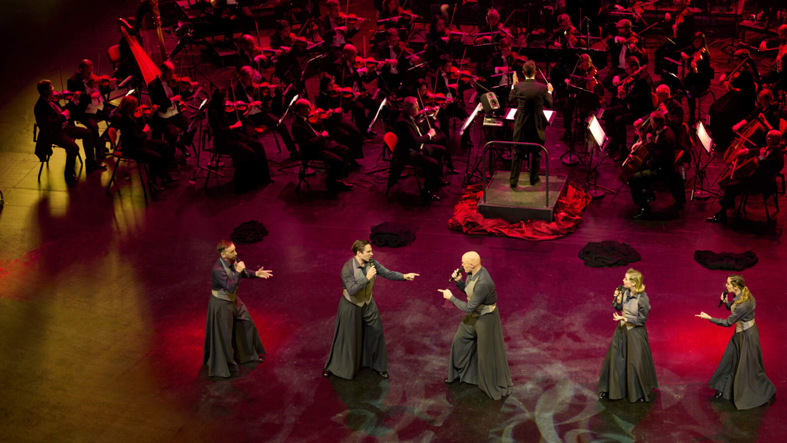 En hel orkester och sex sångare uppträder på en stor scen upplyst i rött och gult ljus. De har fotograferats från ovan.