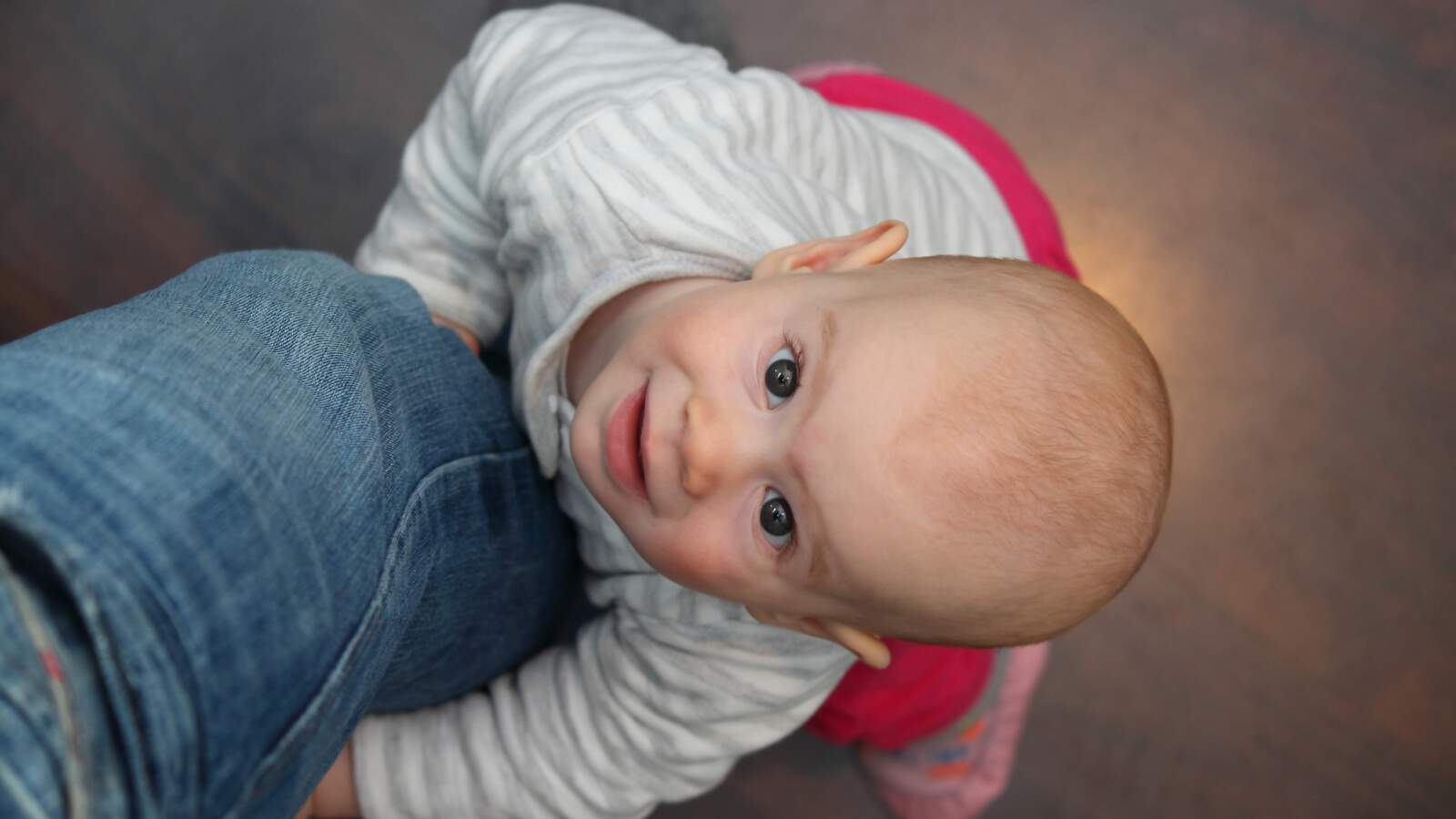 Fotograferad ovanifrån står en ettårig bebis, som håller fast i en vuxens ben och tittar upp mot kameran.