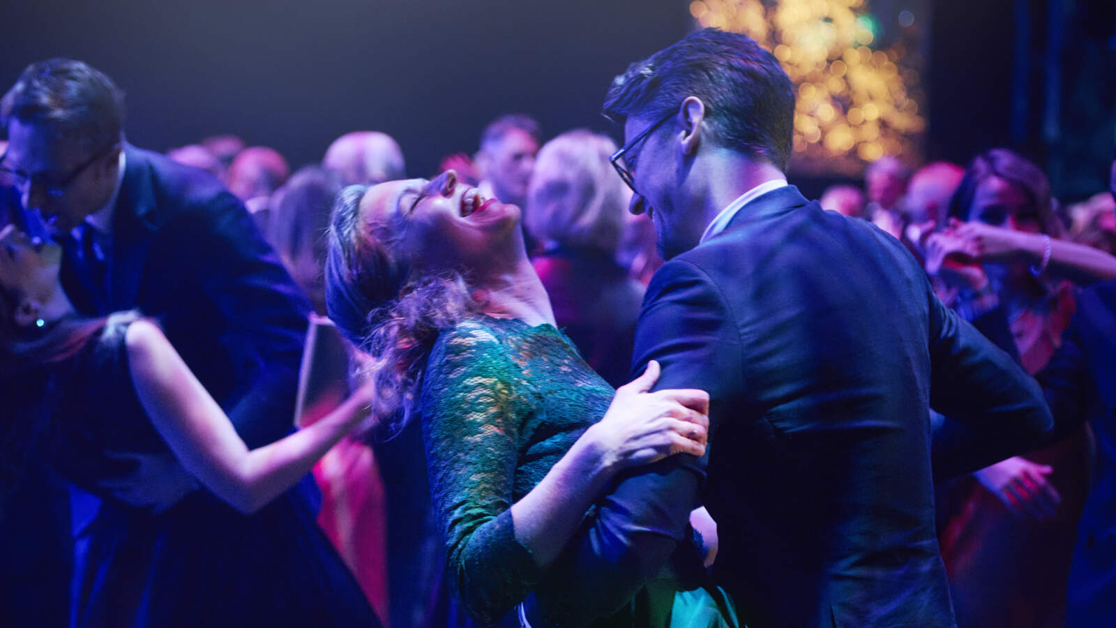 Många par dansar i en balsal under lila ljus. En av paret i förgrunden lutar sig bakåt medan hennes partner håller i henne.