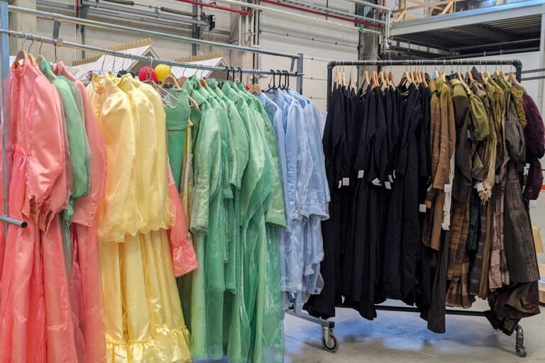 Färgglada teaterkostymer hänger på 2 olika klädställ som står i ett lagerlokal.