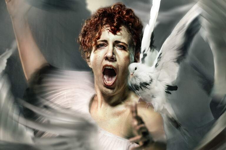 En operasångerska med rött hår står och sjunger bland en flock vita duvor som flyger runt henne.