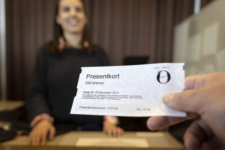 Närbild på en hand som räcker fram ett presentkort till ett leende person bakom disken i en biljettkassa.