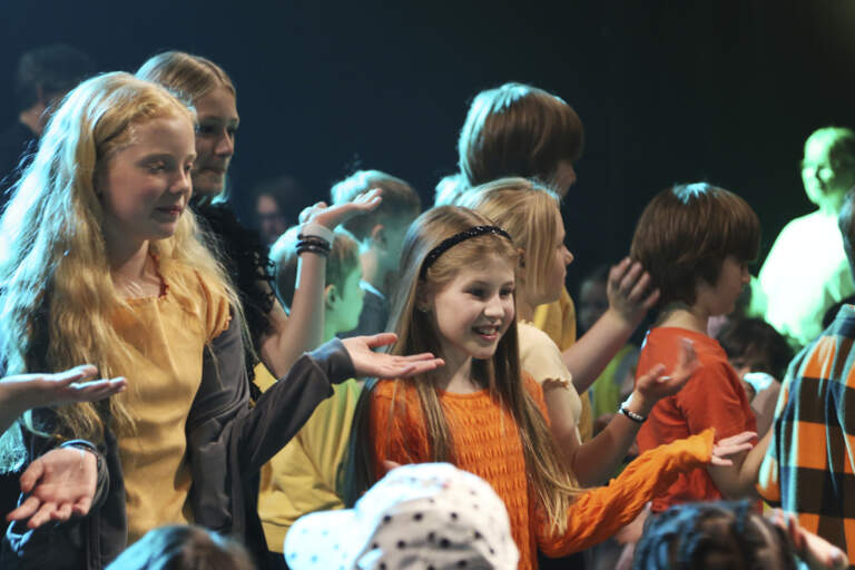 Flera barn ca 8–12 år gamla står med utsträckta händer och sjunger för en grupp yngre barn.