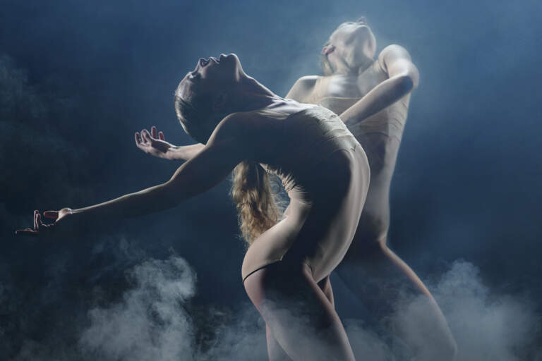 Two dancers stand under white stage lighting in smoke, the dancer in the foreground stands with arms outstretched behind her.
